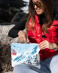 Woman sitting outdoors smiling, wearing red vest and sunglasses, unzipping travel pouch with gondola and snow scene design.