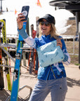 Woman taking a selfie at a ski resort while holding a light blue on blue medium zipper pouch with an all-over skier print.