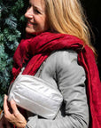 Woman smiling outdoors wearing red scarf and gray jacket, holding silver clutch bag, against greenery background.