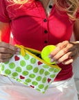 A close-up lifestyle shot of a person holding a white zippered pouch with a green tennis ball pattern and yellow zipper. The pouch is being filled with a bright yellow tennis ball.