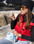 A Woman using a zippered cosmetic pouch featuring a colorful winter mountain landscape print with a red cable car, snowy peaks, pine trees, and skis, shown on a white background.