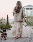Woman in a beige coat and crochet skirt carrying a beige bandana wristlet and walking a dog on a sandy path with a beach house in the background.