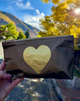 A hand holds a metallic bronze zip pouch with a large gold heart on the front, set against a blurred outdoor background of autumn trees and a mountain.