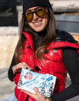 Woman smiling in red vest, sunglasses and hat, holding wristlet with snowy mountain ski design.