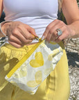 Person holding a yellow pouch with heart designs, wearing a white top and yellow pants.