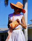 Woman in a colorful outfit with a large orange hat and matching orange bandana pouch against a palm tree-lined street.