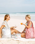 A mother and daughter having picnic on the beach. Mom wearing a silver tote bag on her shoulder and daughter on right wearing pink tote bag.
