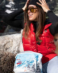 Woman in an outdoor social setting wearing hat, sunglasses and a red vest, holding two zipper pouches with an alpine snowy mountain scene and red gondola print.