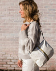 Woman wearing the white shimmer crossbody purse over her shoulder while walking past a brick wall.