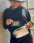 Person holding a card with 'H!' on it, wearing a black outfit, silver fanny pack and a hat, standing indoors.