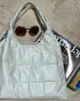 Shimmer white puffer tote bag laying in sand with sunglasses, magazine, and sunscreen