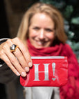 A woman in a red scarf holds up a red card pouch with a large silver letter 'H' and exclamation mark on the front.