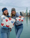 Two women holding bags with heart patterns by a waterfront with a city skyline in the background.
