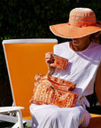 Woman sitting on a chair holding orange bandana patterned bags, wearing an orange sun hat.