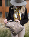 A woman wearing a wide-brimmed taupe hat, dark brown jacket, and a yellow-and-white patterned scarf holds bronze pouches decorated with gold stars, hearts and crescent moons in an outdoor setting.