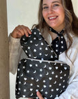 A woman wearing a white lace blouse holds up a black zippered pouch with a white celestial print of stars, moons, and hearts. She is also wearing a matching black neck scarf with the same white pattern.