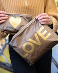 Woman holding a matching set of bronze metallic zipper pouches with oversized gold LOVE lettering and gold heart design in an outdoor autumn setting.