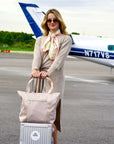 Woman smiling holding luggage and beige tote bag standing outdoors near plane