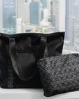 Black signature tote and matching zip pouch set on a bed with a city skyline view behind.