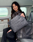 A woman with a scarf sits inside a plane, holding a black signature tote with patterned fabric lining and handles with a matching medium pouch