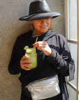 Person wearing a black hat and black outfit with silver fanny pack, holding a green drink with a straw, standing against a neutral wall.