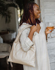 Woman standing holding a glass of wine and shimmer beige puffer tote on shoulder