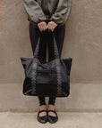 Woman standing on a stoop holding the black signature tote by its straps in front of her.