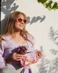 Woman holding a colorful floral pouch and a small plant against a white wall with shadows.