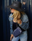 Woman wearing a black hat and denim jacket, holding a bandana patterned wristlet against a dark blue door.