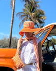Woman wearing an orange hat, orange wristlet and white outfit standing next to a vintage car with palm trees in the background.