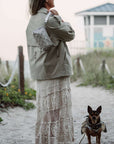 Woman in a beige jacket, beige bandana wristlet and crochet skirt standing on a sandy path with a dog, near a beach house.