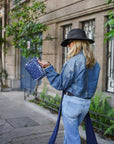 Woman in denim jacket and hat holding a blue bandana bag on a city street.