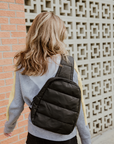 Woman in a gray sweatshirt wearing the black puffer sling backpack across her back beside a brick wall