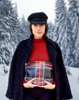 Woman in red top and fur coat holding Medium zipper pouch in tartan plaid pattern against snowy tree background.