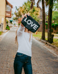 Woman on a cobblestone street holding up a black "LOVE" pouch on a tree-lined lane.