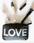 Black "LOVE" pouch flat lay spilling out makeup essentials — foundation, lipstick, brush, and skincare products.