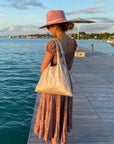 Woman in pink hat and dress standing on ocean dock with shimmer beige shoulder tote