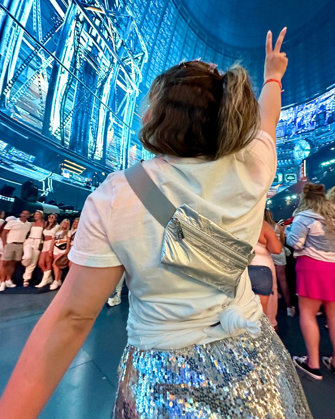A person seen from behind wearing a white t-shirt, a silver metallic crossbody bag, and a silver sequined skirt, standing in a large indoor venue for a concert with blue lighting and other people in the background.