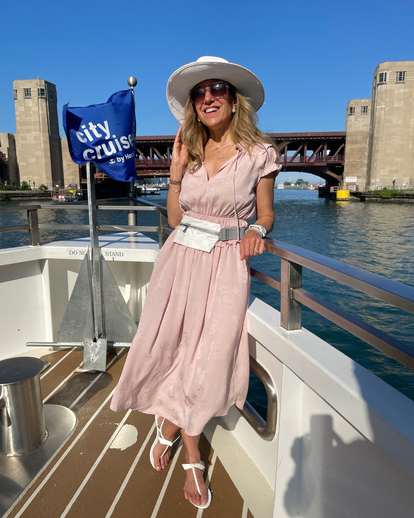 A woman posing on a boat wearing a light pink midi dress with a white belt, a white sun hat, and white sandals, with a blue 'city cruise by the bay' flag and the Chicago River North area in the background.