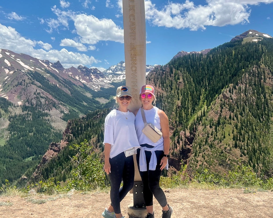 Two people standing next to a mountain sign with a scenic mountain landscape in the background.