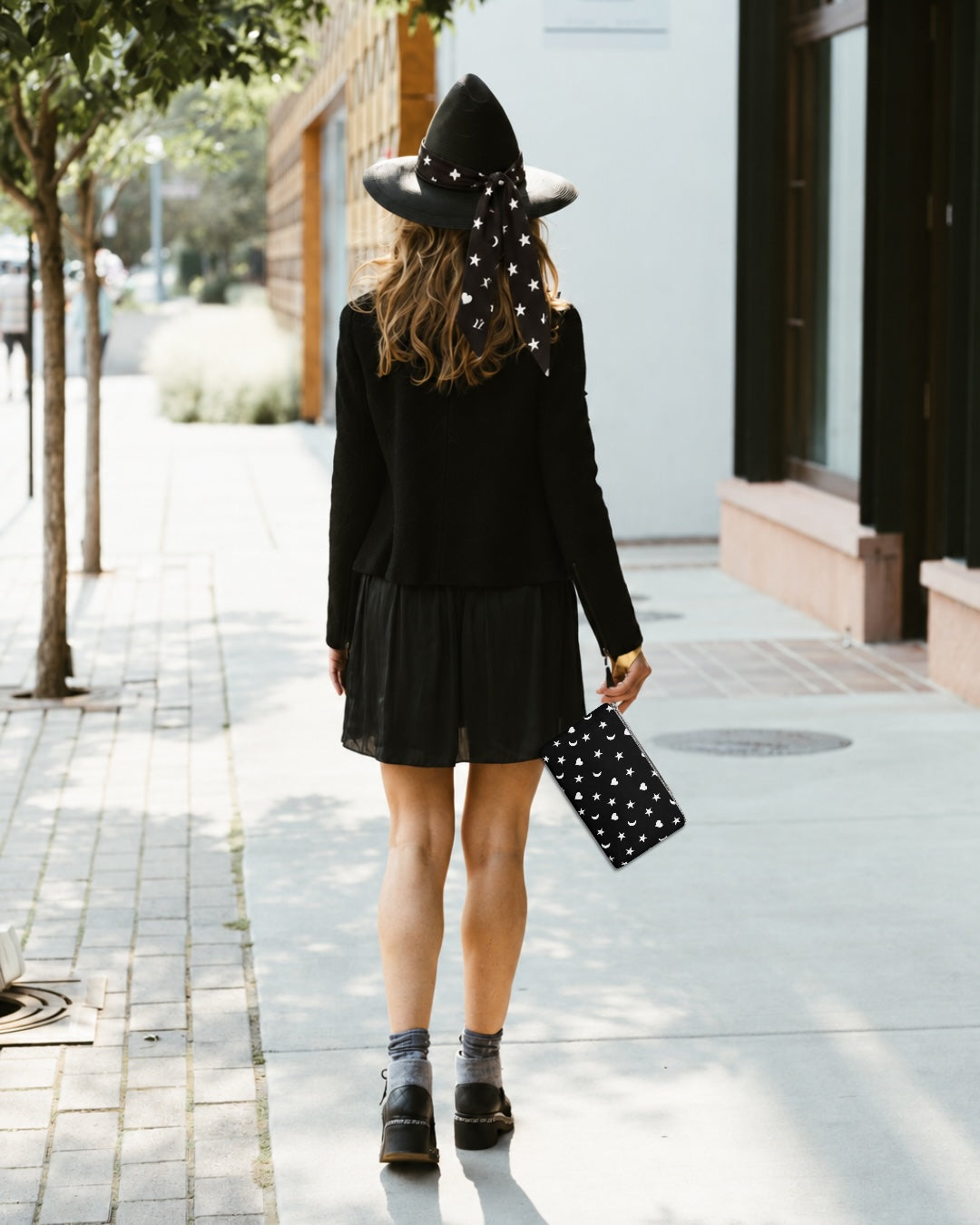 Women walking wearing all black holding a black heart moon and star wristlet and wearing a black hat with a scarfette wrapped around it.
