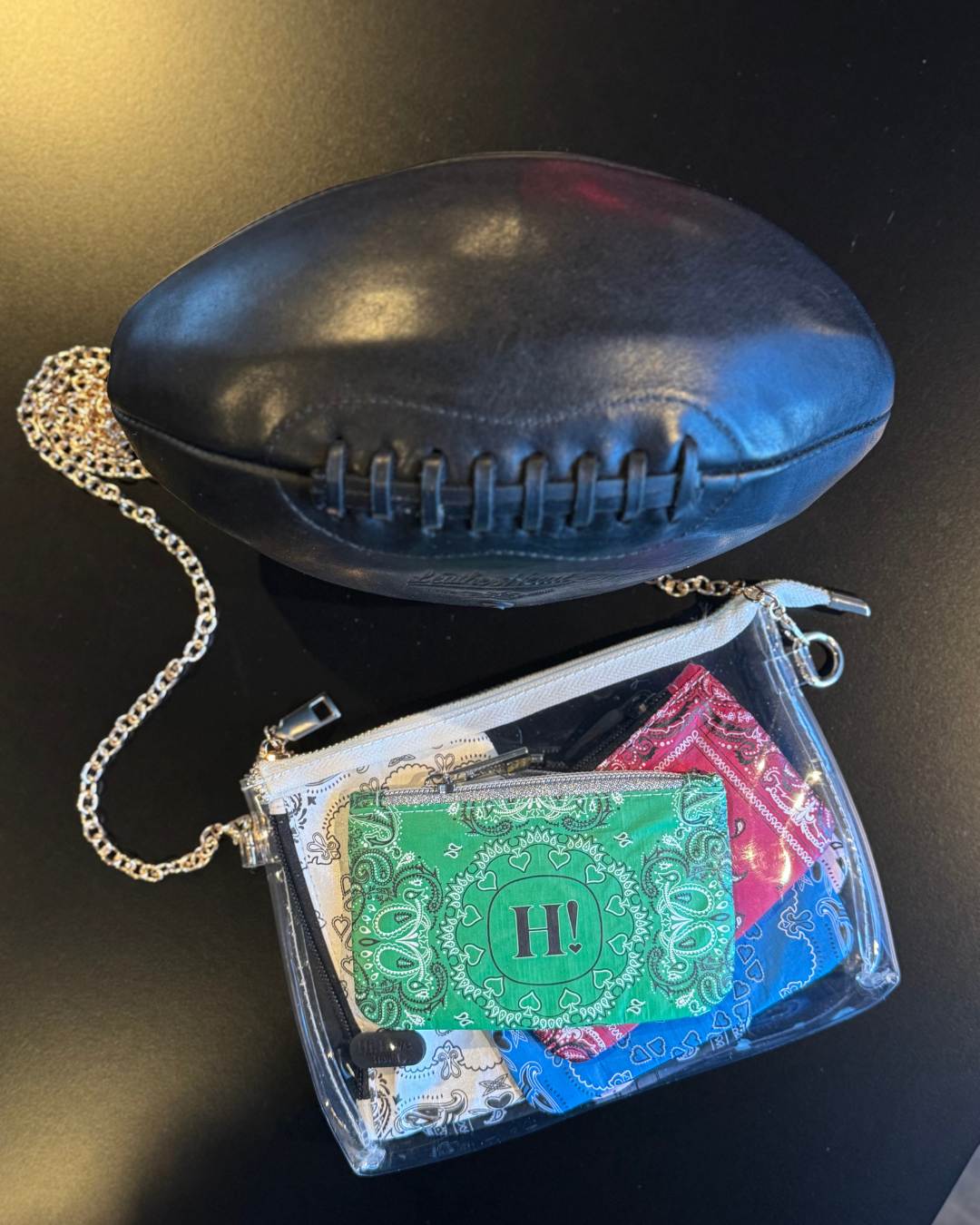 A black leather football is shown next to a clear zippered pouch containing colorful bandana-print card pouches inside, on a dark background.