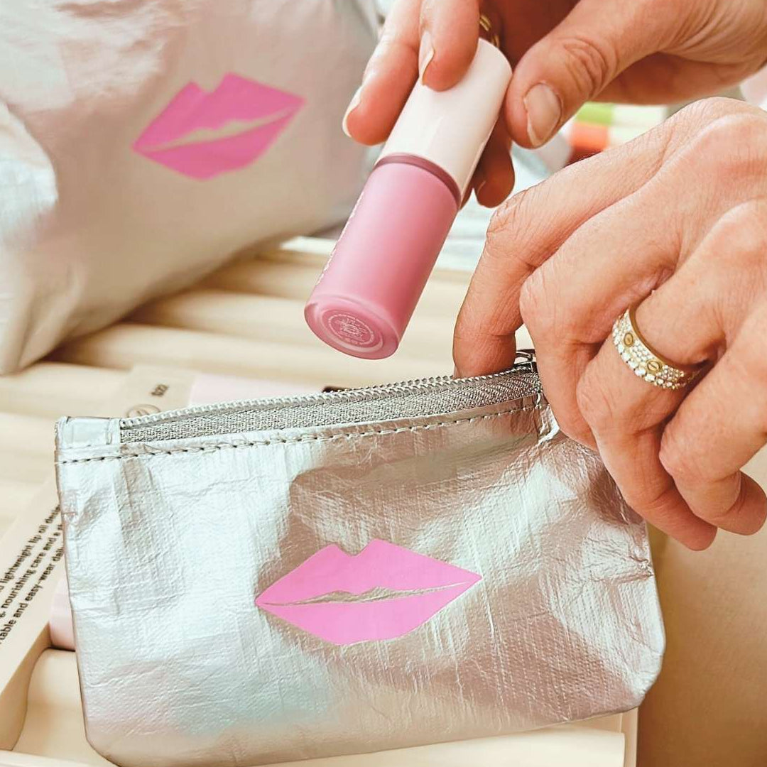 Person placing lip gloss in a silver makeup bag with pink lip design.