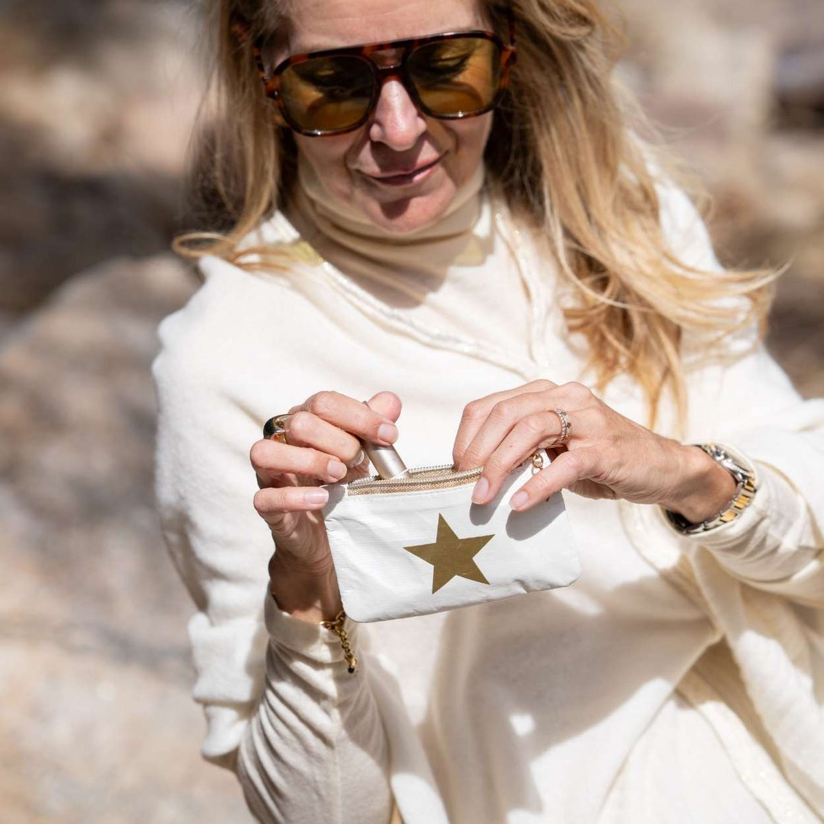 A woman wearing a cream turtleneck sweater and tortoiseshell sunglasses holds a small white pouch with a gold star in a sunny outdoor setting.