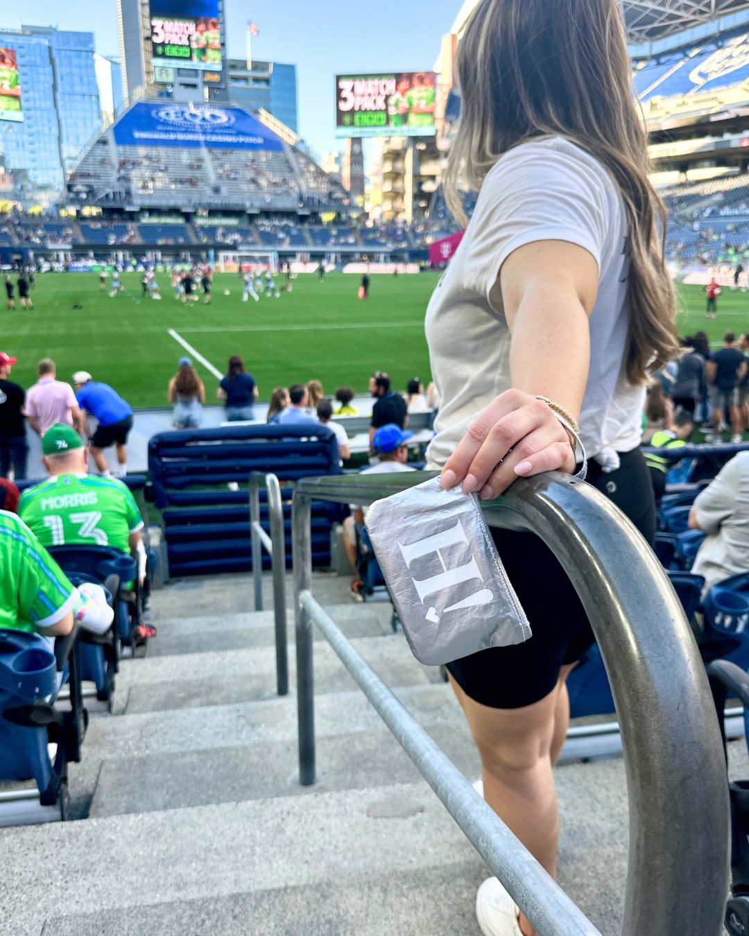 A woman in a stadium stands on concrete stairs holding a silver card pouch with a white 'Hi' logo, with a soccer game in progress in the background.