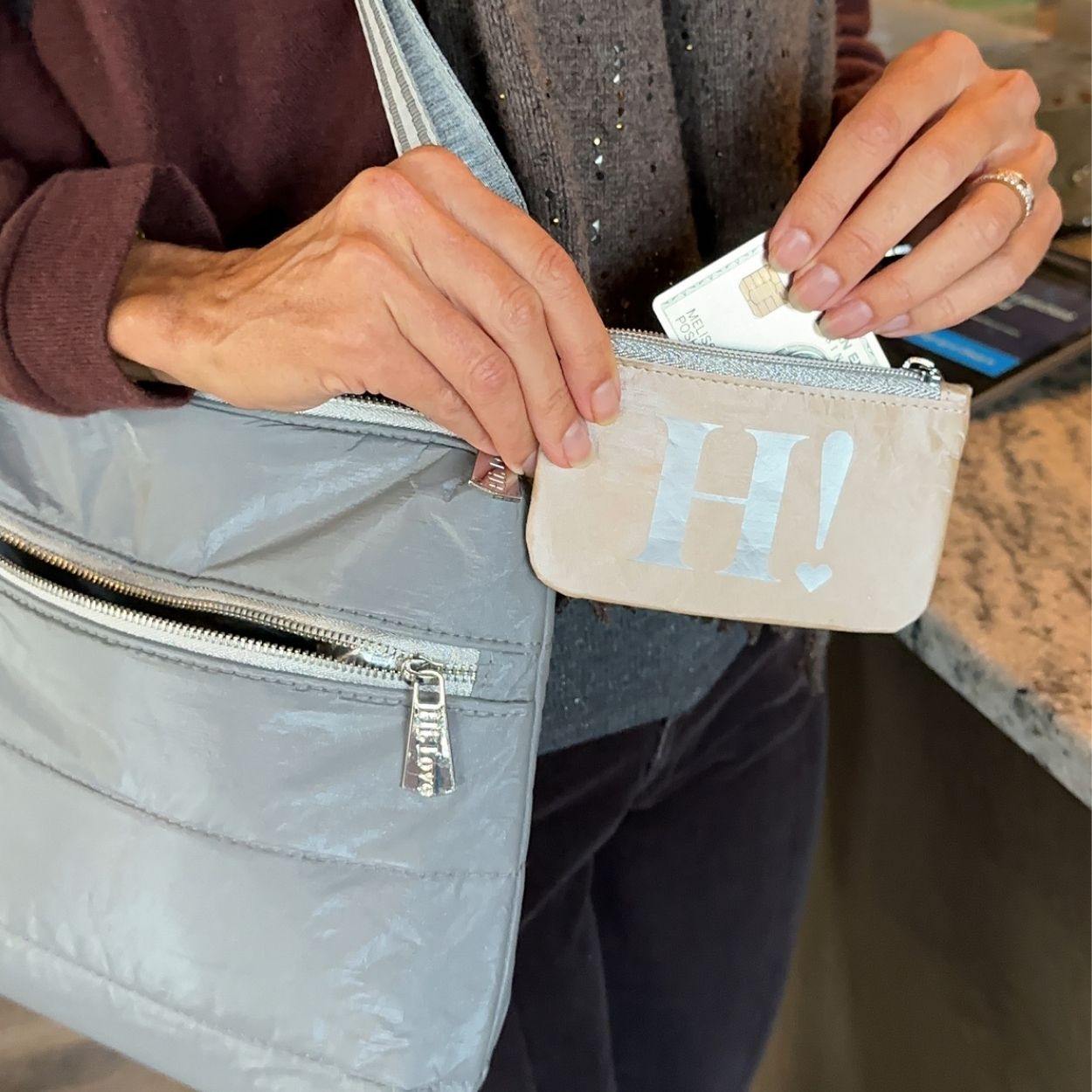 A close-up lifestyle shot of a person holding a light gray quilted shoulder bag and reaching into a small beige coin pouch with a silver 'H!' logo. The person is wearing a brown sweater and dark pants, and the scene is set in an indoor environment with a marble countertop in the background.