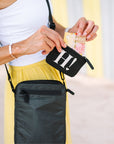 A woman wearing a black crossbody phone bag and a smaller black zip pouch with a white 'HI!' print, holding the pouch open to show money inside. The background is a soft, blurred yellow and white architectural setting.