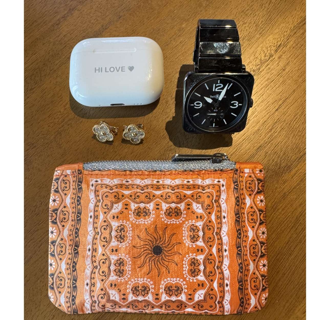 Orange Bandana pattern clutch with black watch, ear buds, and earrings on a wooden surface