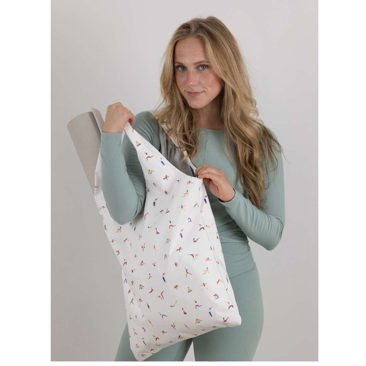 Woman in green athletic apparel holding a yoga pose print tote bag with yoga mat inside.