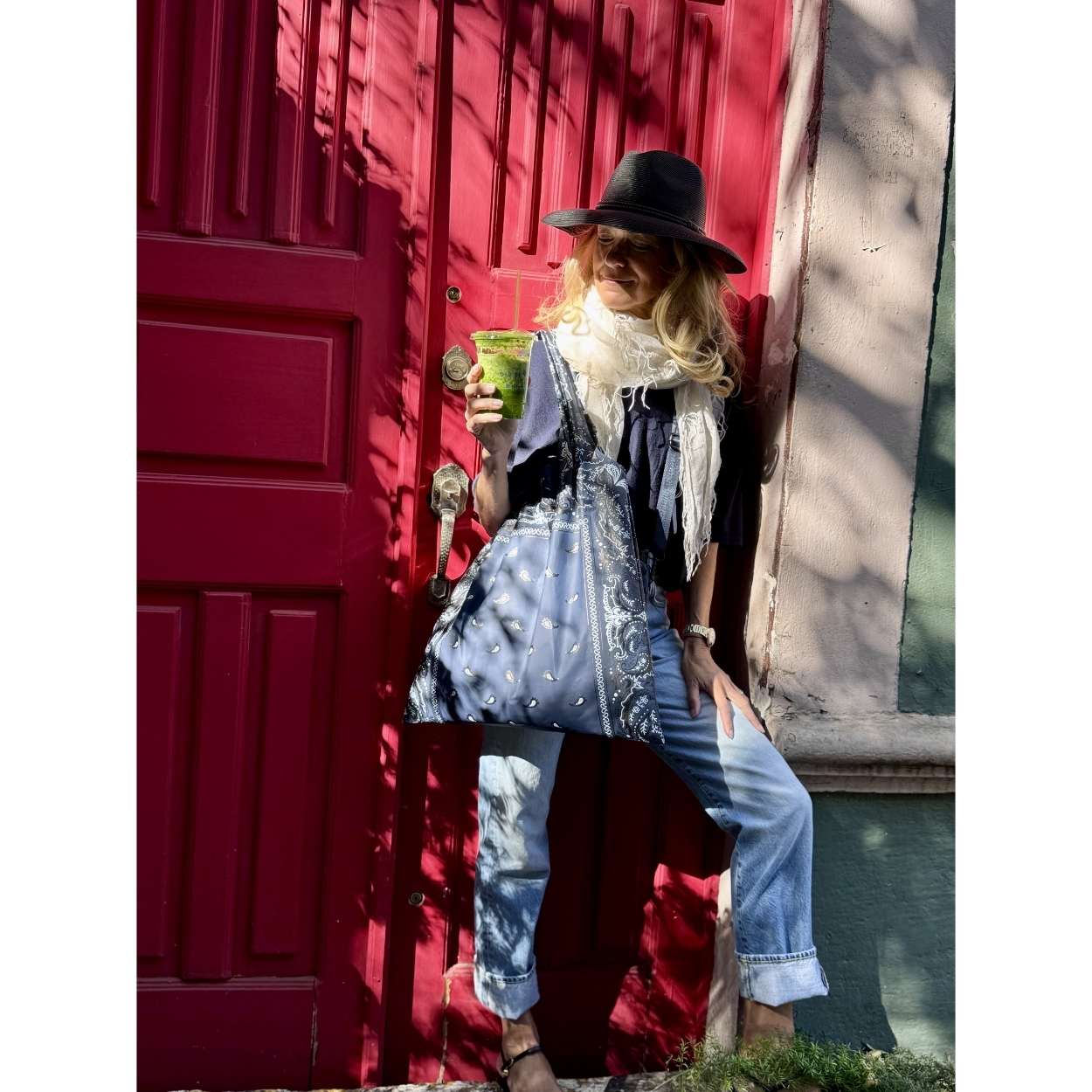 Person carrying navy bandana print tote bag leaning against a red door holding a green cup, wearing a black hat, scarf, and blue jeans.